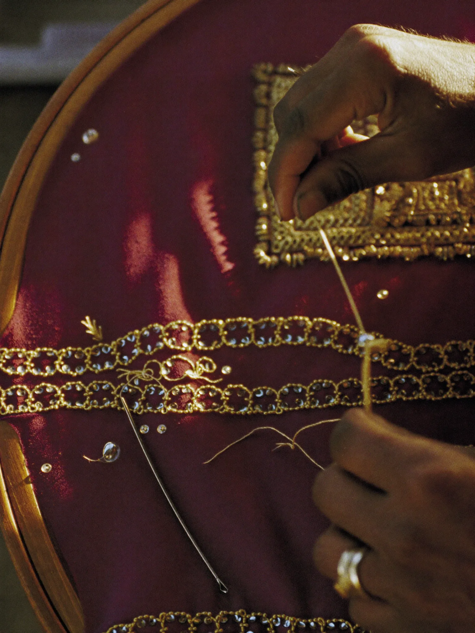 Two hands couching gold thread onto oxblood velvet stretched on a wooden karigar's frame