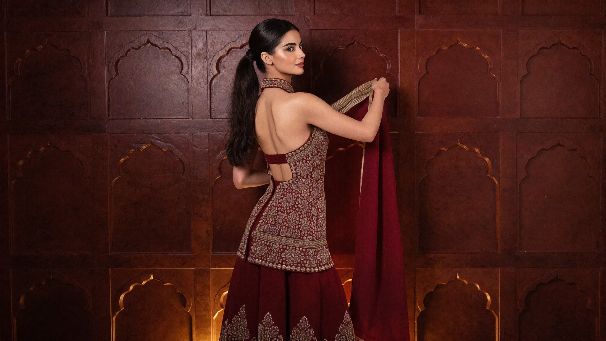 A maroon sharara worked in silver and gold, held against a carved sandstone wall of Mughal arches