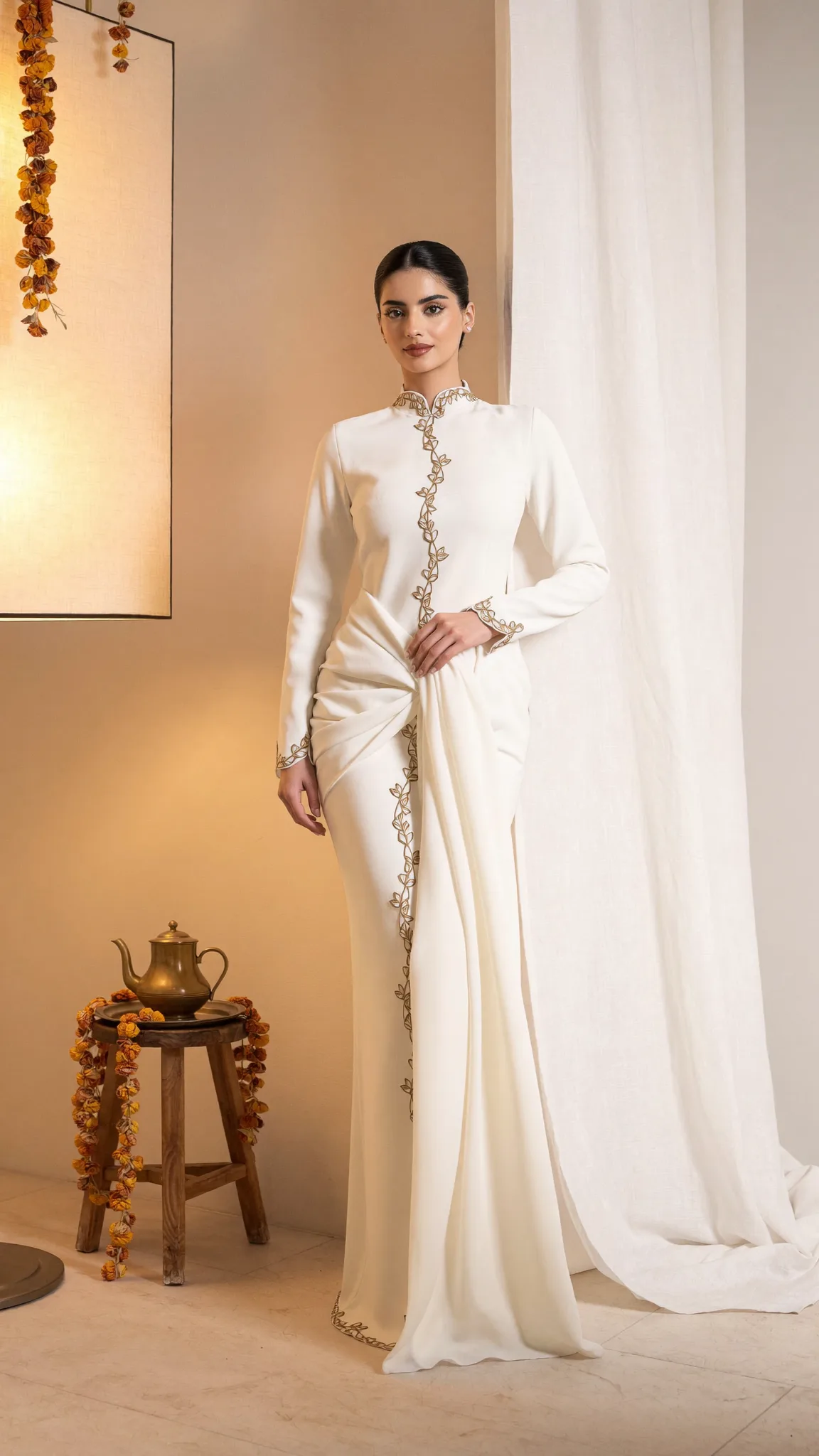 An ivory kurta and drape with a gold vine placket, beside marigold garlands and a brass pot