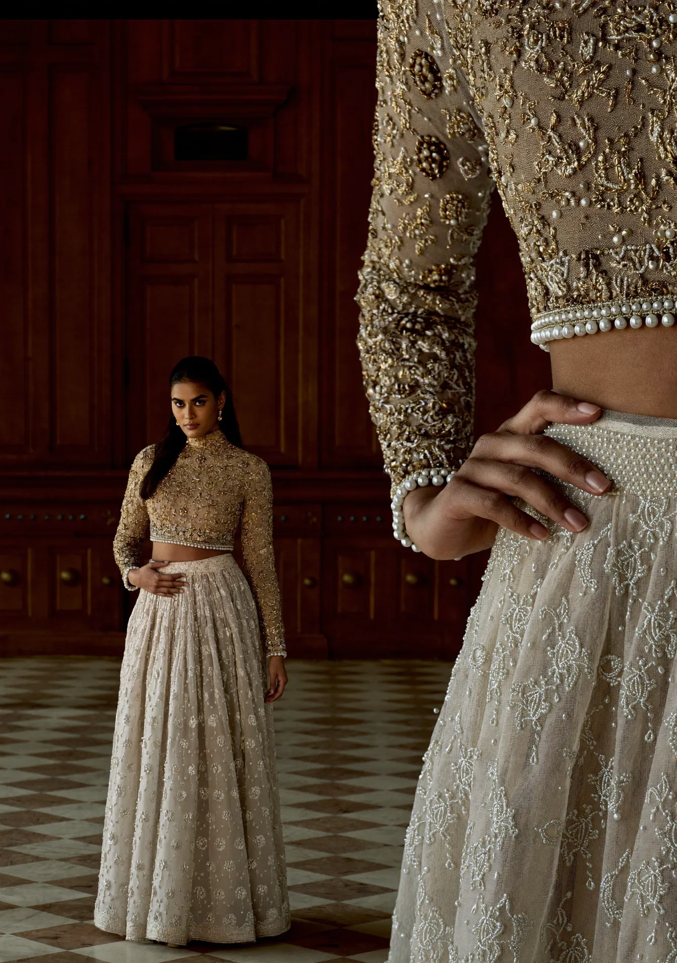 Two looks in gold and pearl in a panelled hall, one close at frame right, one far across a checkerboard floor