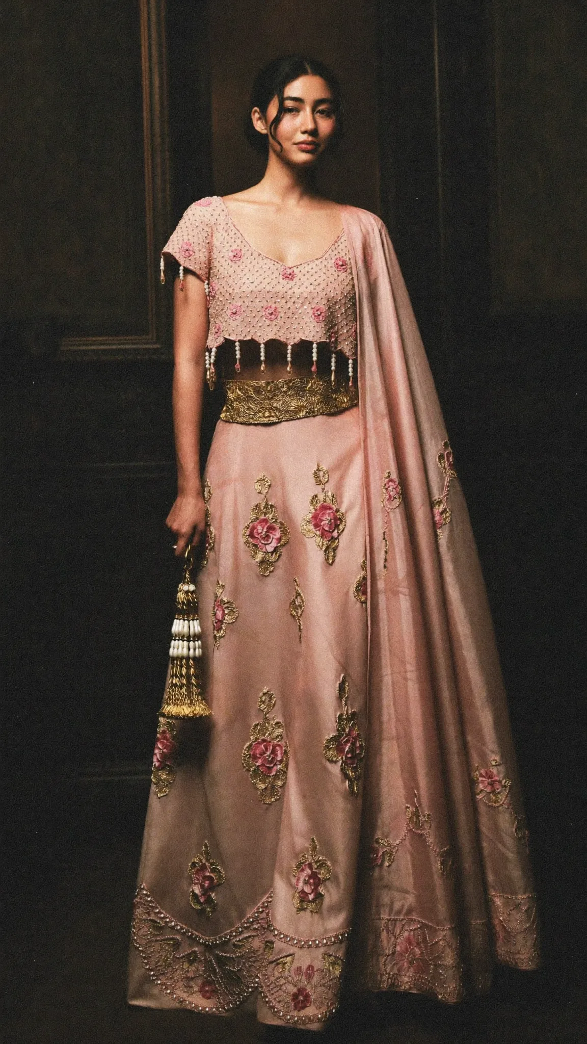 A blush lehenga worked in raised rose appliqué with a gold belt and pearl tassel