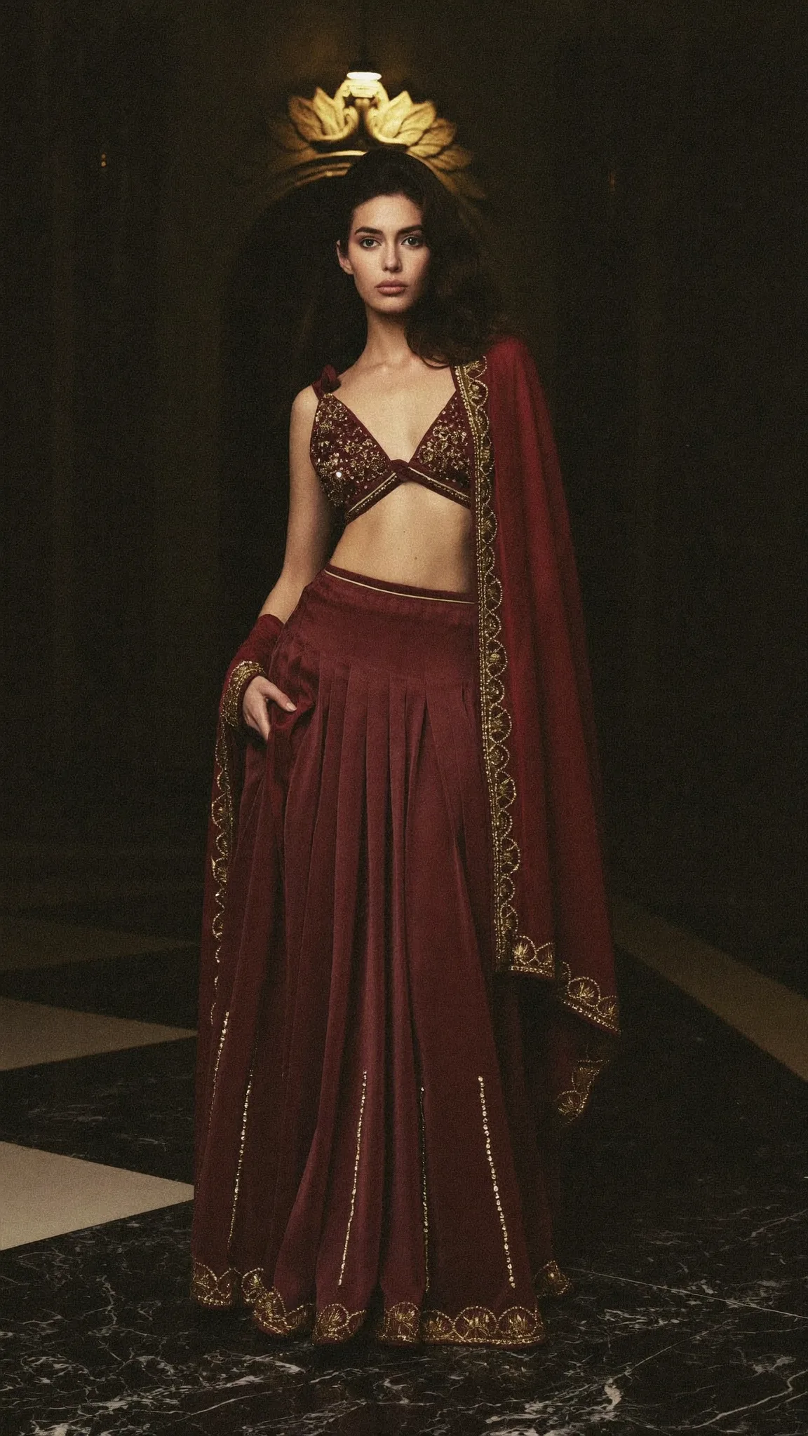 An oxblood velvet lehenga with a scalloped gold border, on black marble under a gold sconce